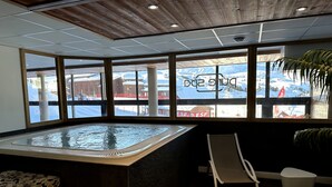 Un espace spa comprenant un bain à remous, des sièges et une vue sur un paysage montagneux enneigé, visible à travers de grandes fenêtres.
