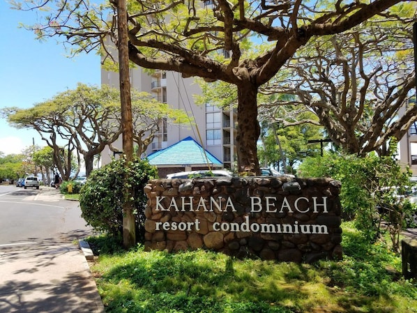 A sign for KAHANA BEACH resort condominium with a stone wall and greenery.