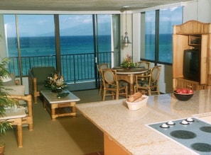 A balcony with a view of the ocean, a dining area with chairs and a table, and a kitchen with a stove and a bowl of fruit.