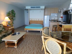 A compact hotel room with a bed, a small kitchenette, a dining area with a glass table, a sofa, and a lamp.