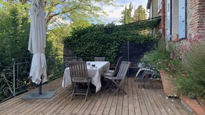 Une terrasse en bois avec une table et des chaises, entourée de verdure et d’un mur de briques.