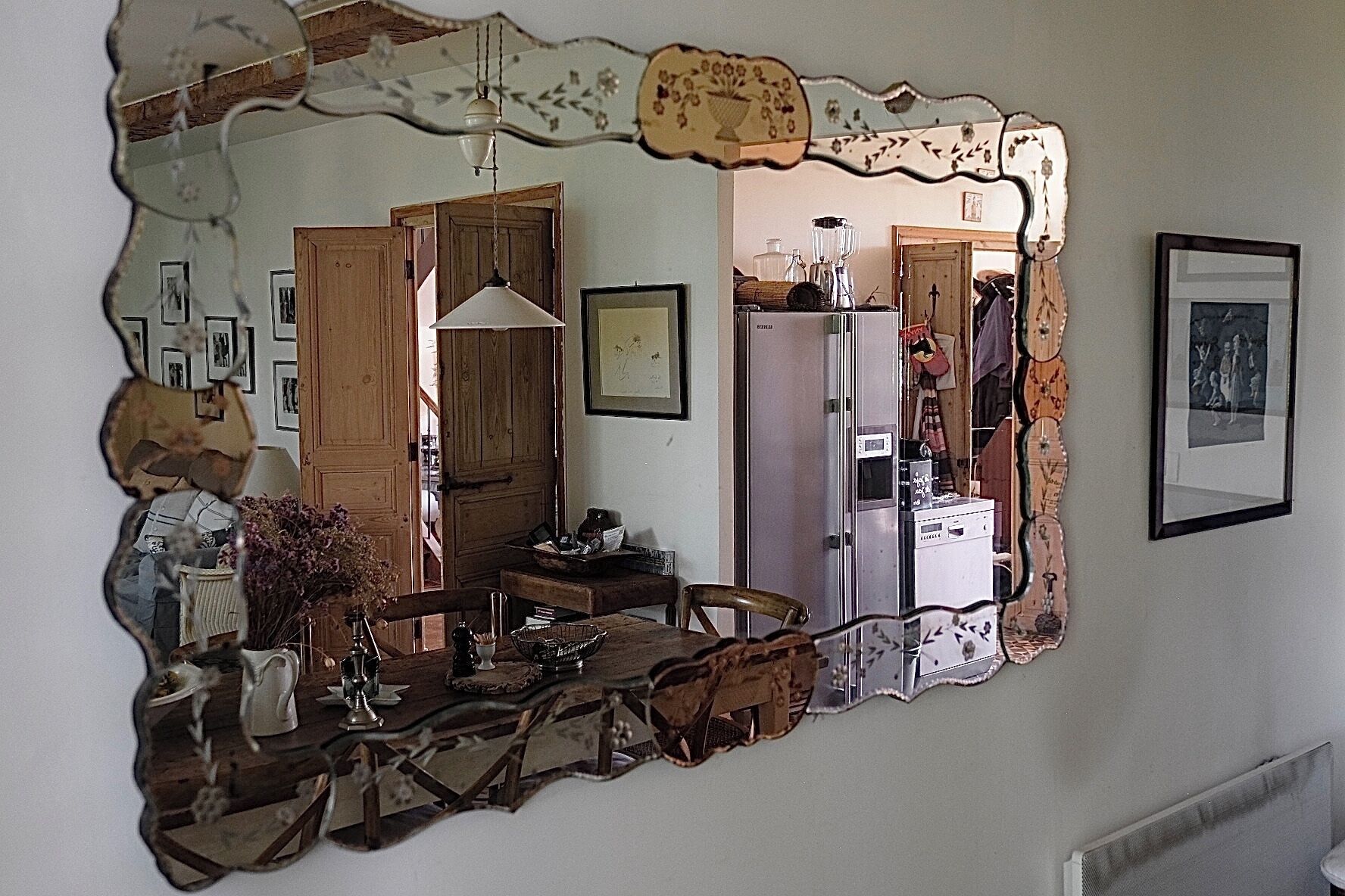 Un miroir reflétant une pièce avec une table à manger en bois, des chaises, un luminaire suspendu et un réfrigérateur.
