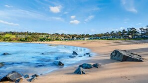 A beach with clear water, rocks, and houses in the background.