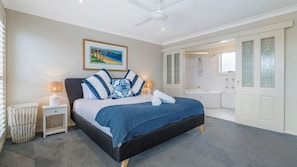 A bedroom with a bed, bedside tables, a ceiling fan, a painting, and a bathroom with a bathtub and shower.