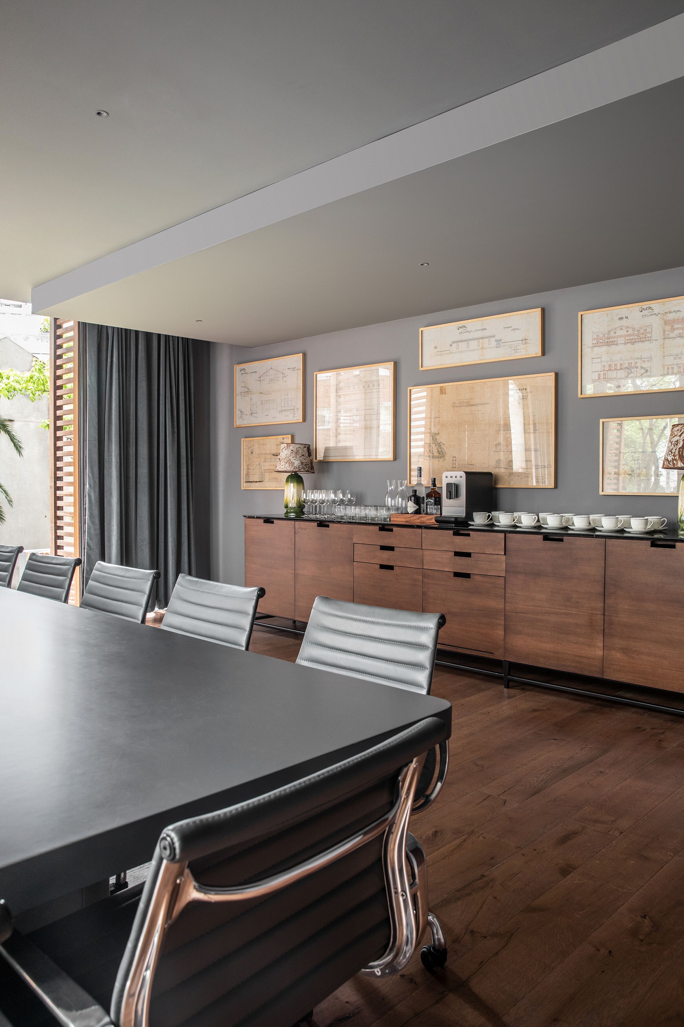 A modern meeting room with a long table, chairs, a coffee station, and framed architectural drawings on the wall.