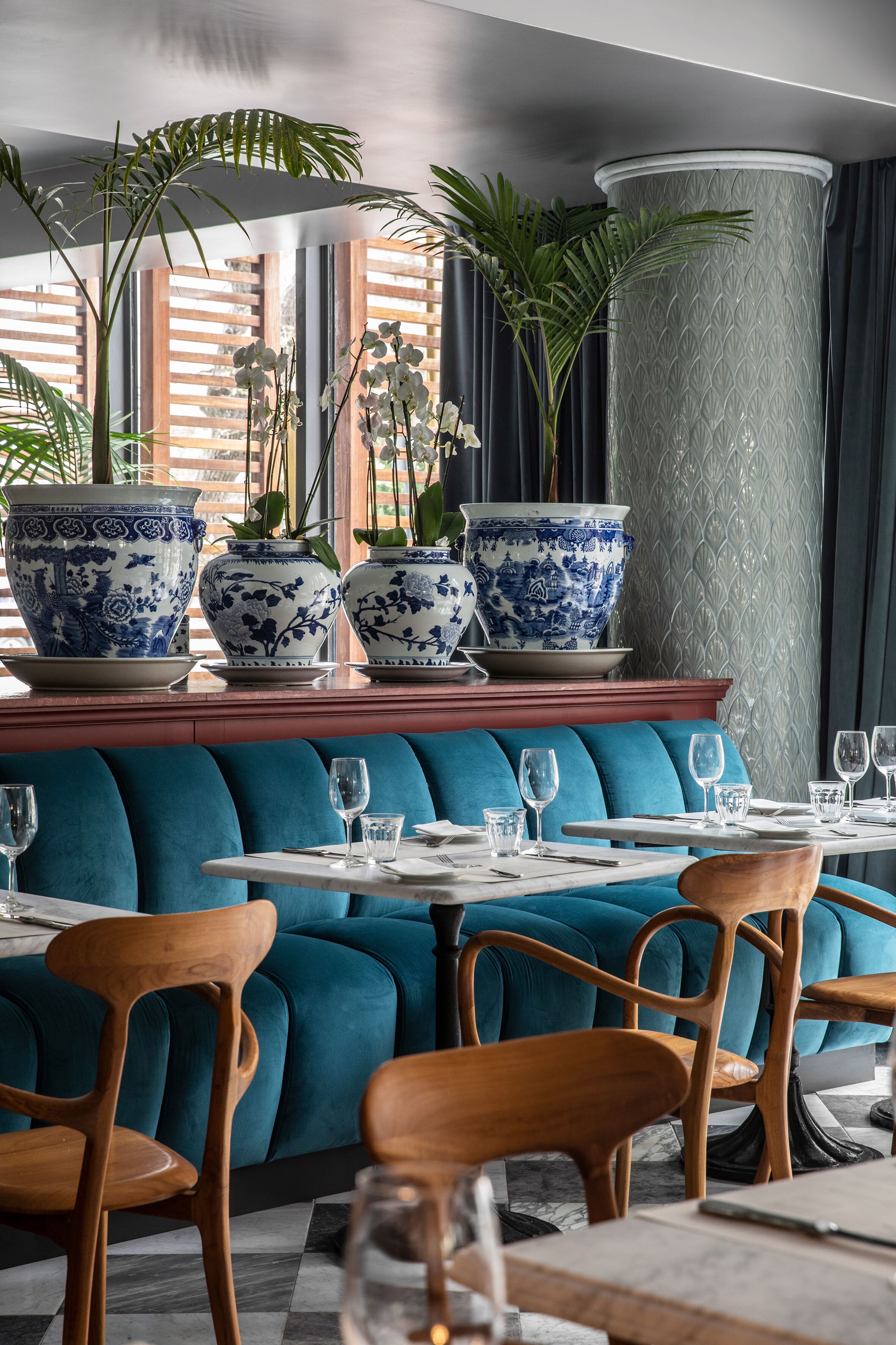 Interior de restaurante con asientos tapizados en azul, sillas de madera y grandes jarrones azules y blancos sobre una mesa central.