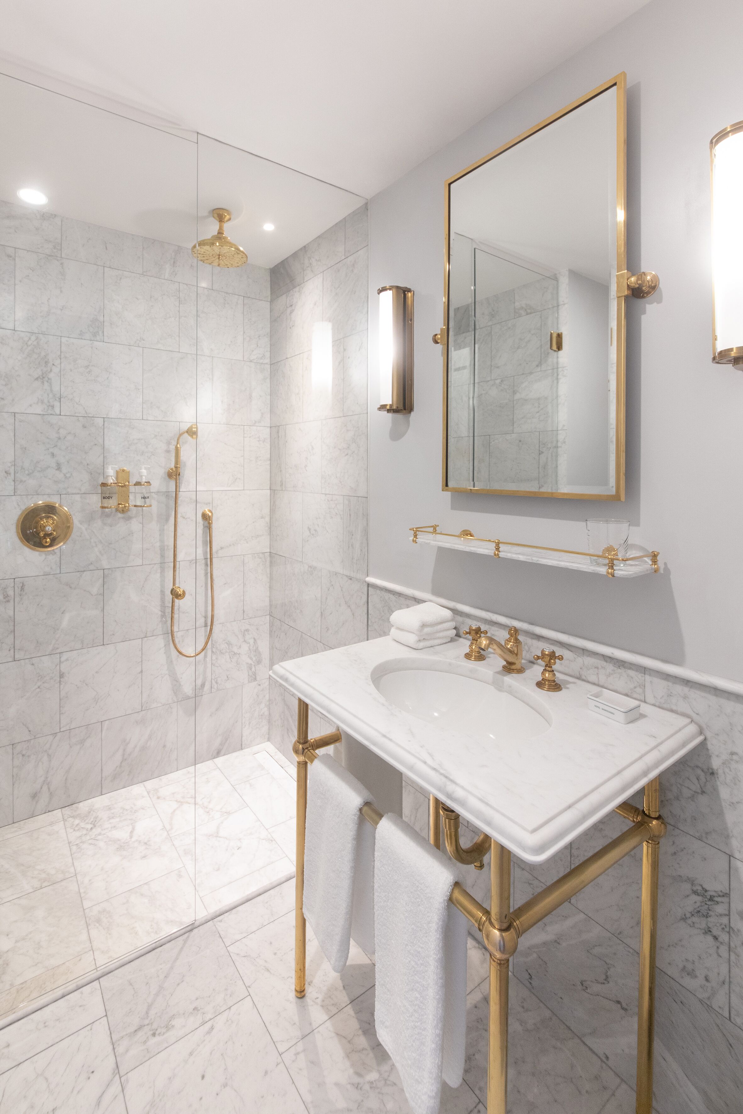 Baño con lavamanos blanco, detalles dorados, cabina de ducha de vidrio y un espejo grande.