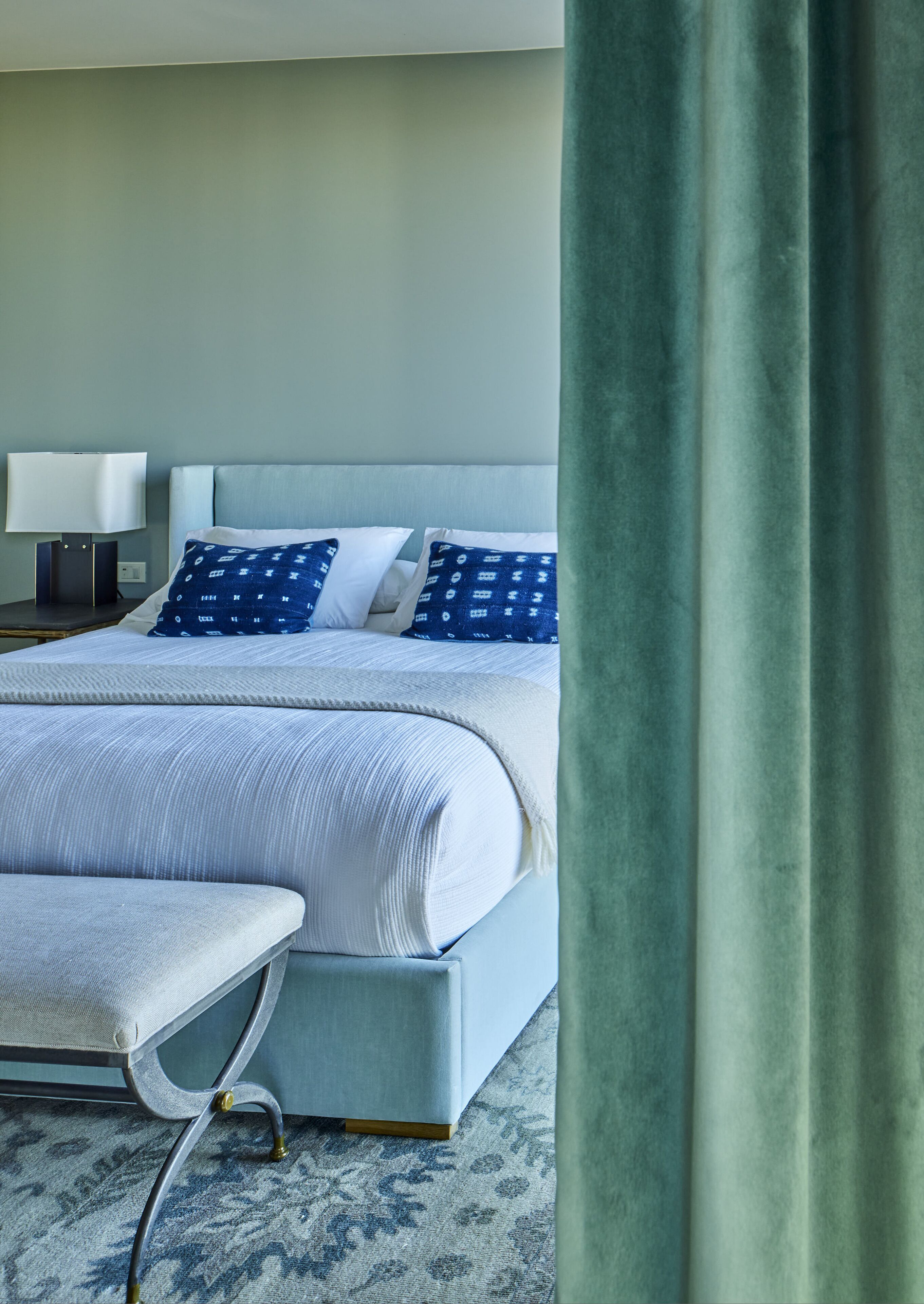 Una cama bien hecha con un edredón blanco y azul estampado, una mesita de noche con una lámpara y un banco al pie de la cama.
