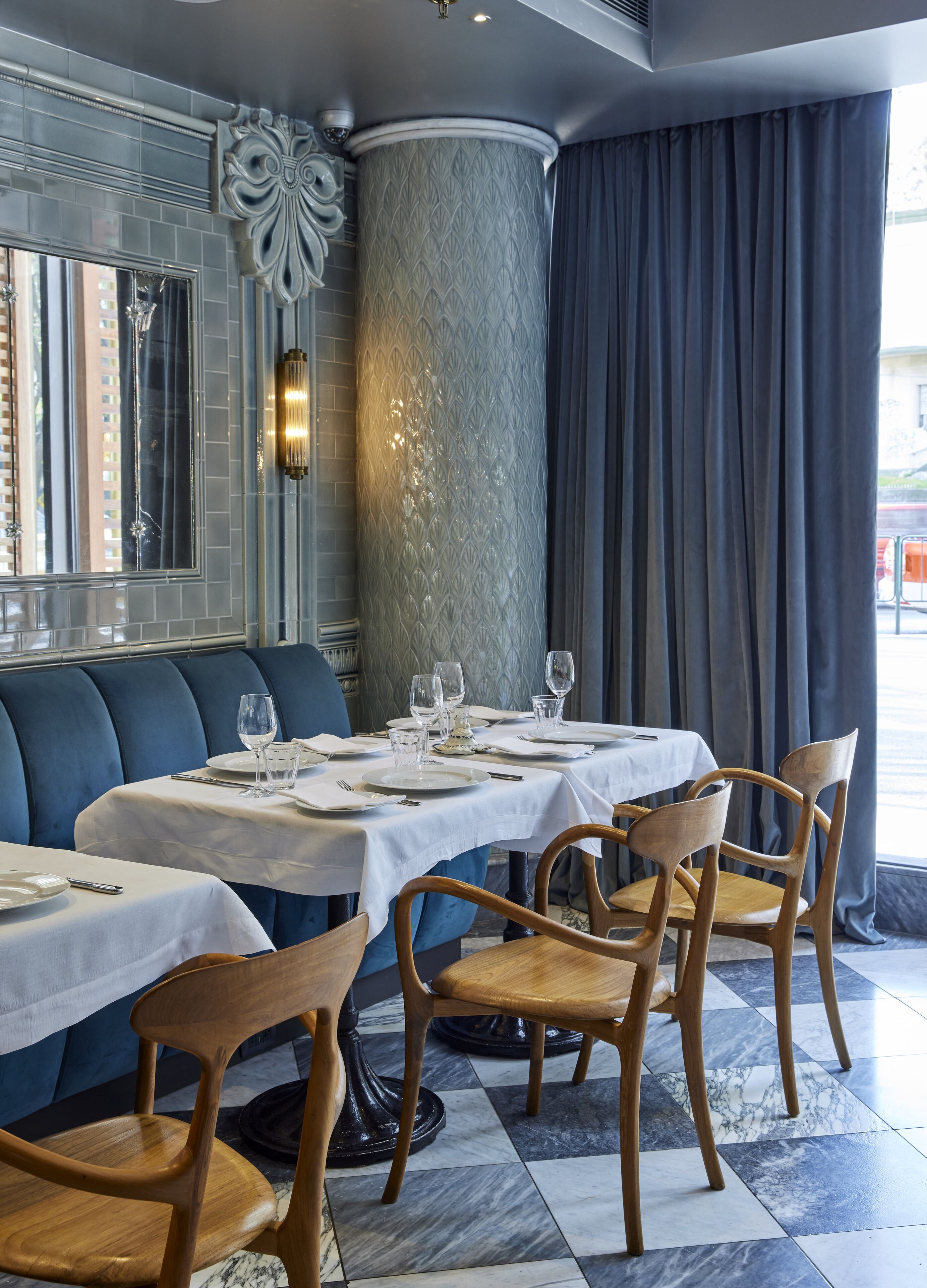 Un restaurante con suelo de baldosas de mármol, asientos tapizados en azul y sillas de madera. Las mesas están dispuestas con manteles blancos y copas de vino vacías.
