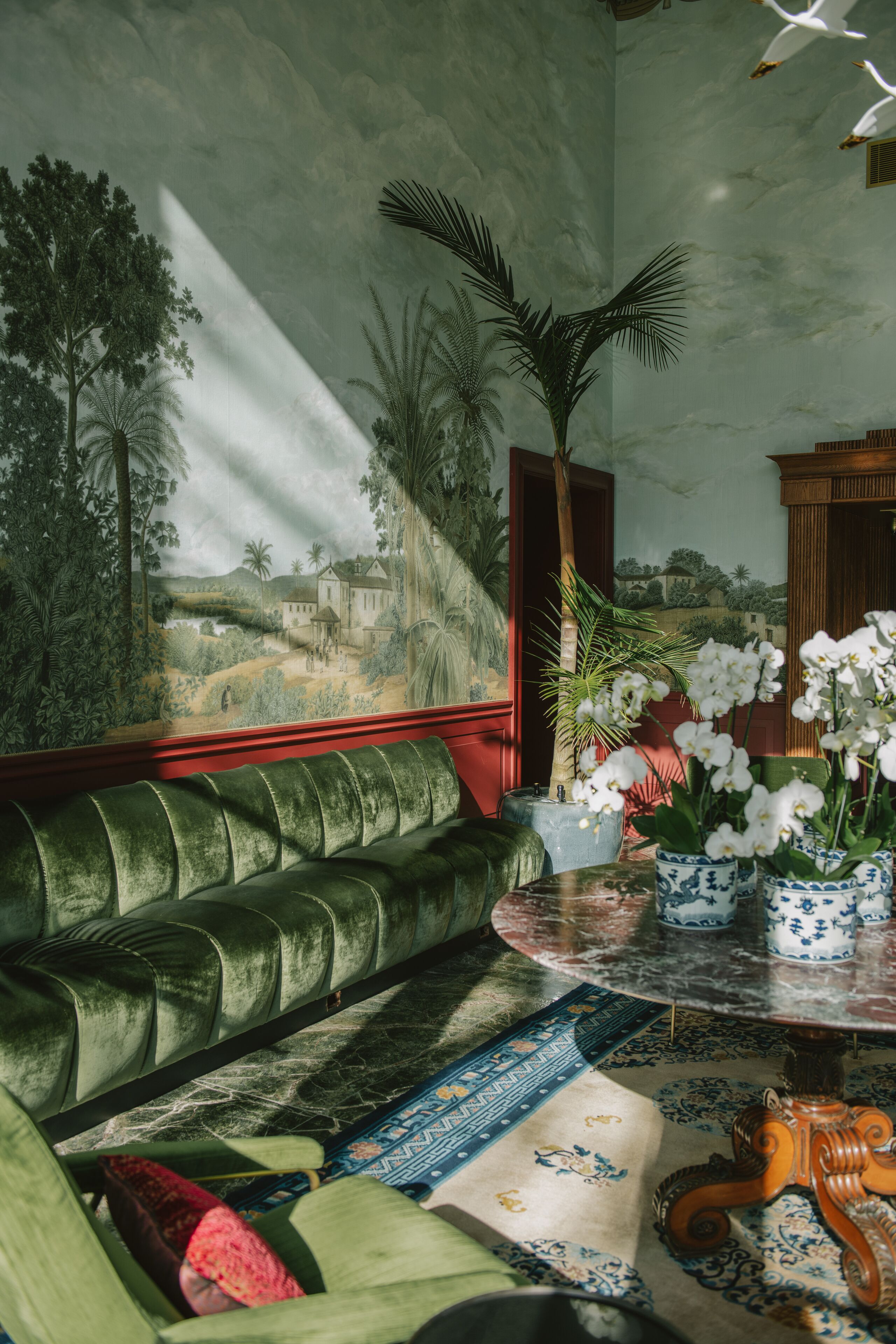 A room with a green velvet sofa, a round table with a vase of white flowers, and a mural of a landscape on the wall.