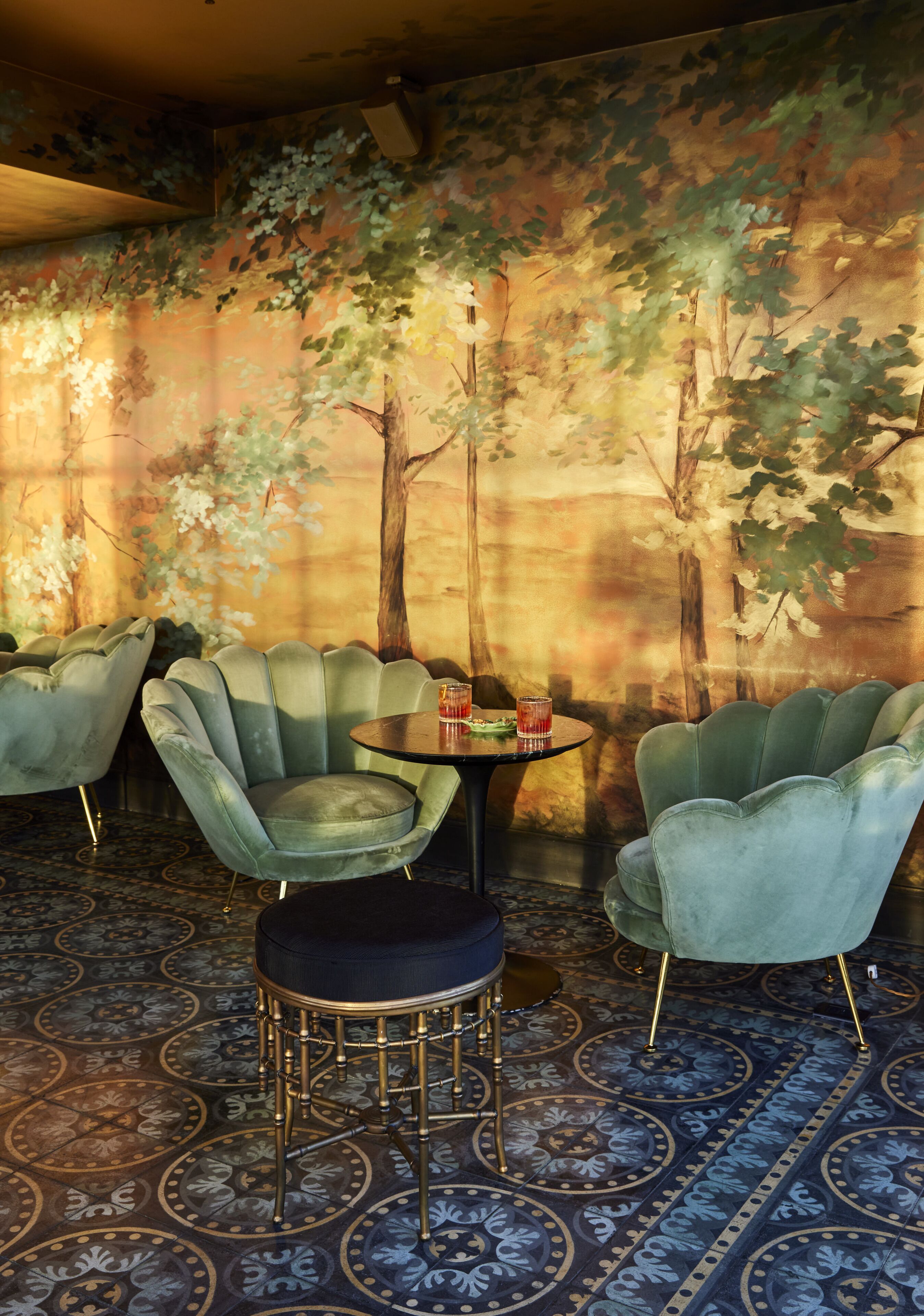 Une pièce dont un mur est décoré d’une fresque représentant des arbres et dont le sol est recouvert d’un motif. On y trouve deux fauteuils verts et une petite table sur laquelle est posée une boisson.