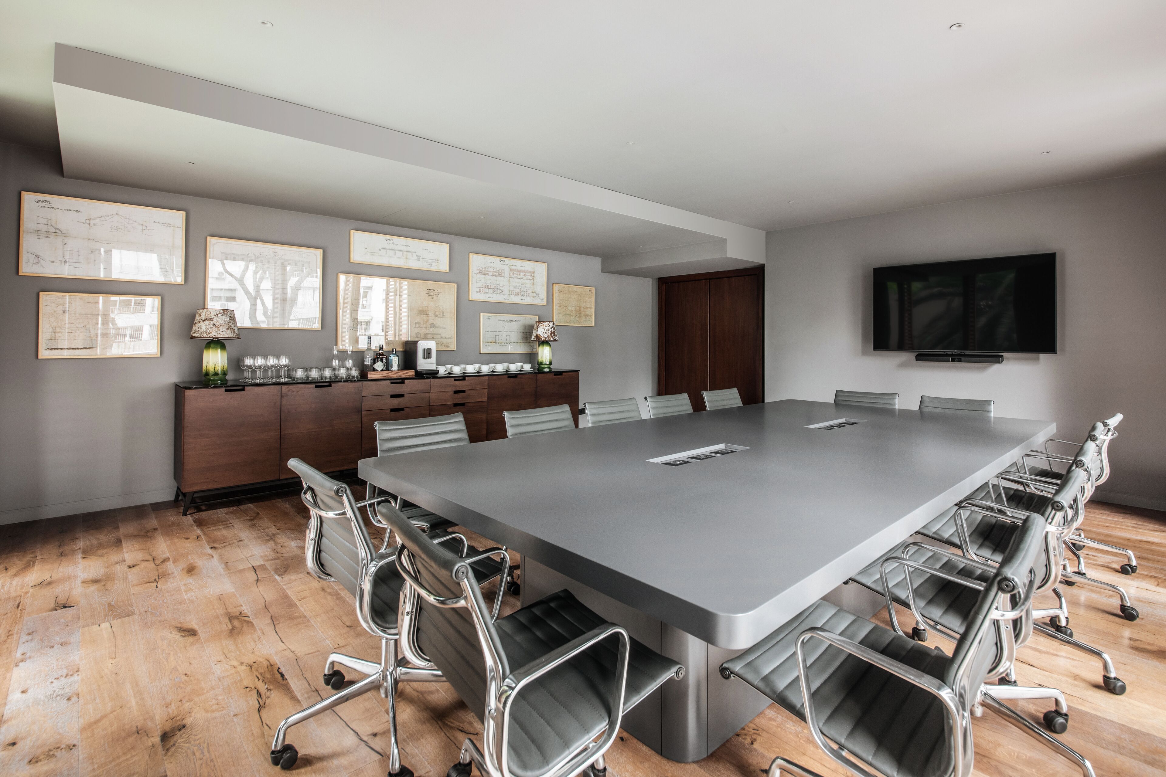 A conference room with a long table, multiple chairs, framed architectural drawings on the wall, a television mounted on the wall, and a wooden floor.