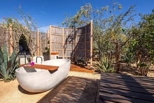 A modern outdoor bathroom with a large bathtub, wooden deck, and surrounding vegetation.