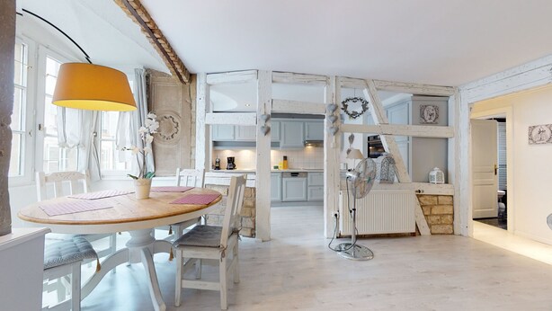 Ein Zimmer mit Esstisch, Stühlen, einer Lampe, einem Heizkörper und einer Küche im Hintergrund.