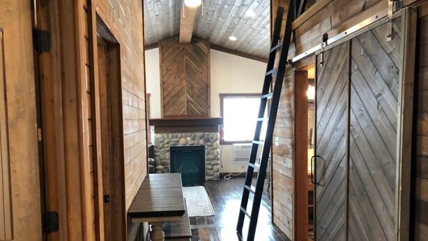 A rustic interior with wooden walls, a stone fireplace, a wooden table, and a ladder.