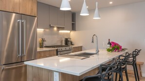 A modern kitchen with stainless steel appliances, a white countertop, and bar stools. - Elegant Condo with Private BBQ and Ocean Views (Saanich North)