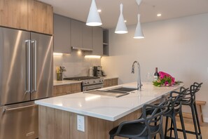 A modern kitchen with stainless steel appliances, a white countertop, and bar stools. - Elegant Condo with Private BBQ and Ocean Views (Saanich North)