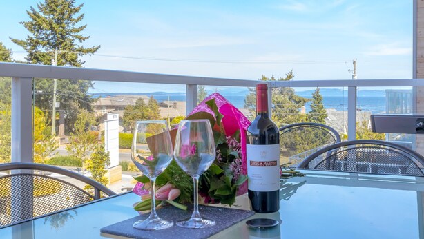 A table with two wine glasses, a bottle of wine, and a bouquet of flowers on a balcony overlooking a scenic view. - Elegant Condo with Private BBQ and Ocean Views (Saanich North)