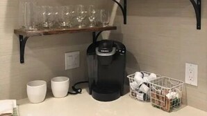 Una cocina con dos estantes metálicos negros que sostienen platos, vasos y tazas, una cafetera y una cesta con objetos sobre la encimera.