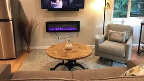 Una sala de estar con un televisor de pantalla plana montado en la pared, una chimenea debajo, una mesa de centro de madera, un sofá y un sillón con cojín.