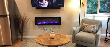 Ein Wohnzimmer mit Flachbildfernseher an der Wand, darunter ein Kamin, ein Couchtisch aus Holz, eine Couch und ein Sessel mit Kissen.