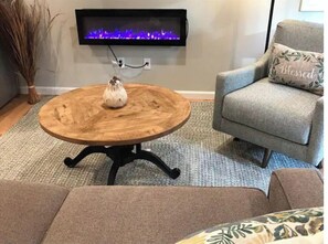 Ein runder Couchtisch aus Holz mit einer weißen Vase, ein graues Sofa mit einem Kissen mit der Aufschrift 'Blessed' und ein an der Wand befestigtes Fernsehgerät mit violetten Flammenanzeigen.