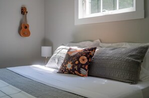 A bed with white bedding, a floral pillow, and a grey quilt. A ukulele is mounted on the wall to the left. - Dog Friendly • Hot Tub • Firepit • Wine Country (Fennville)