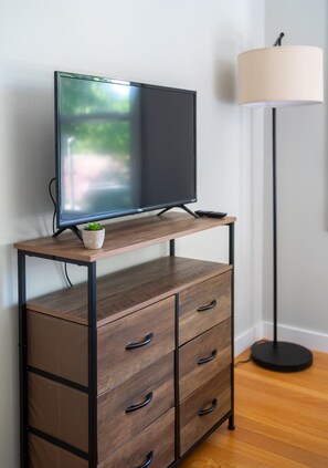 Een flatscreen televisie op een houten tv-meubel met lades, een kleine plant en een vloerlamp.
