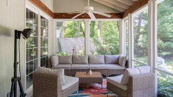 A covered outdoor seating area with a ceiling fan, wicker furniture, and a view of greenery.