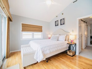 Um quarto com uma cama, mesas de cabeceira, uma janela com persiana e uma porta para outro cômodo.