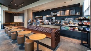 Un café moderno con un largo banco de cuero marrón, mesas redondas y una barra que exhibe comida y bebidas.