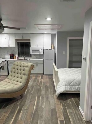Cozy retreat with a compact kitchen, microwave, and adjacent seating area with a tufted armchair and a bed.