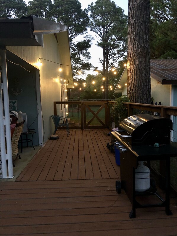 Una terraza de madera con una parrilla, luces colgantes y una zona de estar cubierta.