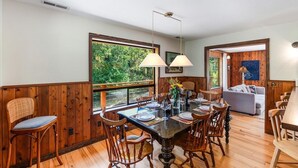 Un coin repas avec une table en bois dressée pour quatre personnes, entourée de chaises en bois, une grande fenêtre donnant sur un paysage verdoyant et un luminaire suspendu.