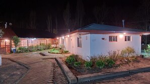 Edificio pequeño de una sola planta con techo rojo y fachada blanca. Tiene un camino de entrada pavimentado y un pequeño jardín con diversas plantas.