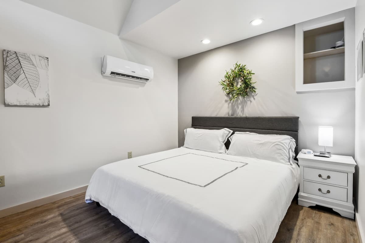 Quarto moderno com uma cama grande, roupa de cama branca, mesa de cabeceira com abajur e ar condicionado na parede.