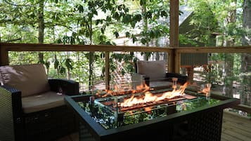 Une terrasse extérieure dotée d’un foyer à flammes sous une table en verre, de meubles en osier et offrant une vue sur une zone boisée.