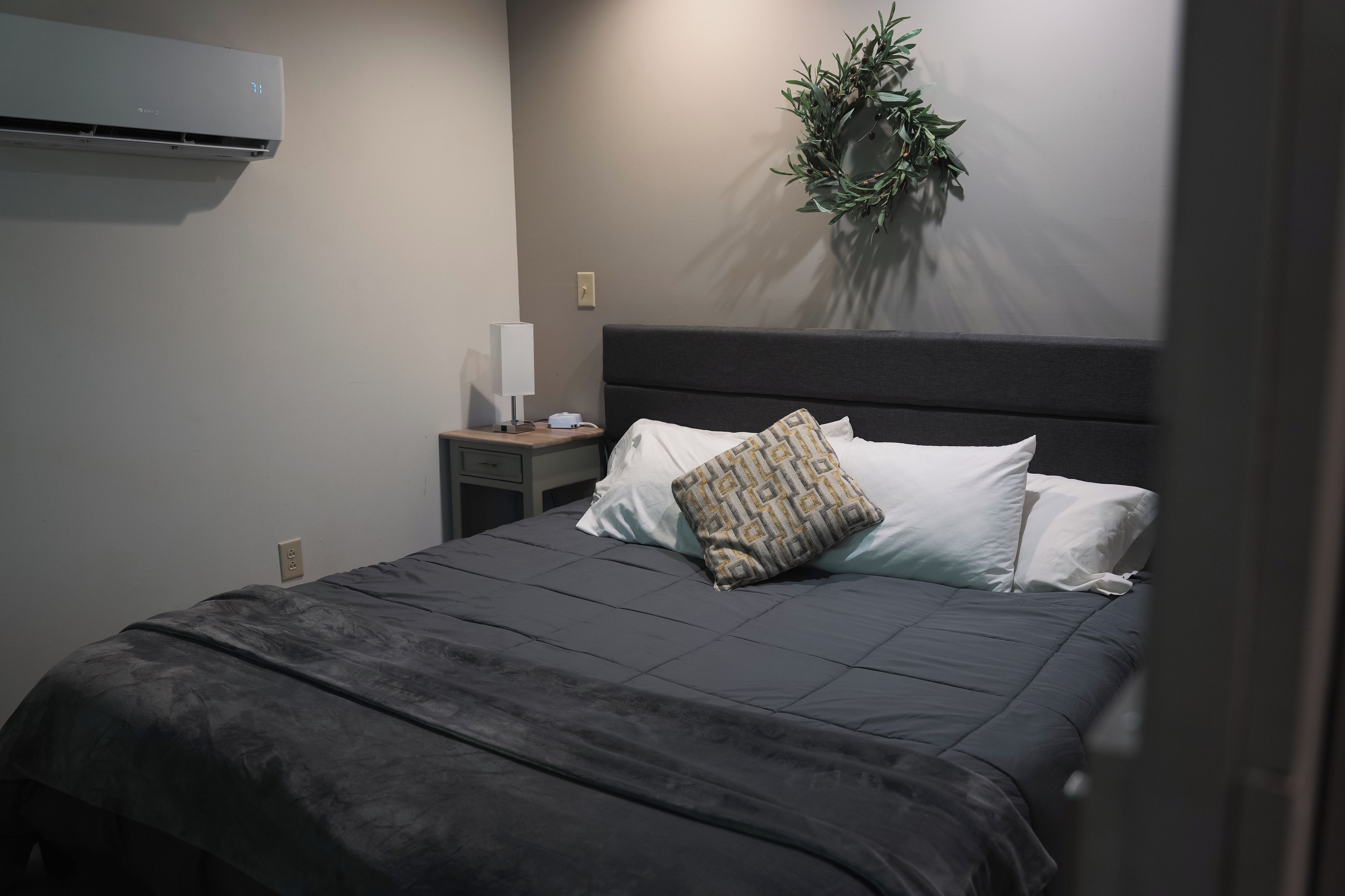 Una cama bien hecha con una colcha gris oscuro y almohadas, una mesita de noche con una lámpara y un enchufe, y un aire acondicionado de pared.