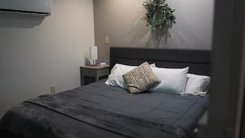Una cama bien hecha con una colcha gris oscuro y almohadas, una mesita de noche con una lámpara y un enchufe, y un aire acondicionado de pared.