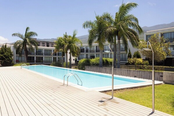 A swimming pool with a wooden deck, surrounded by palm trees and a building with balconies.