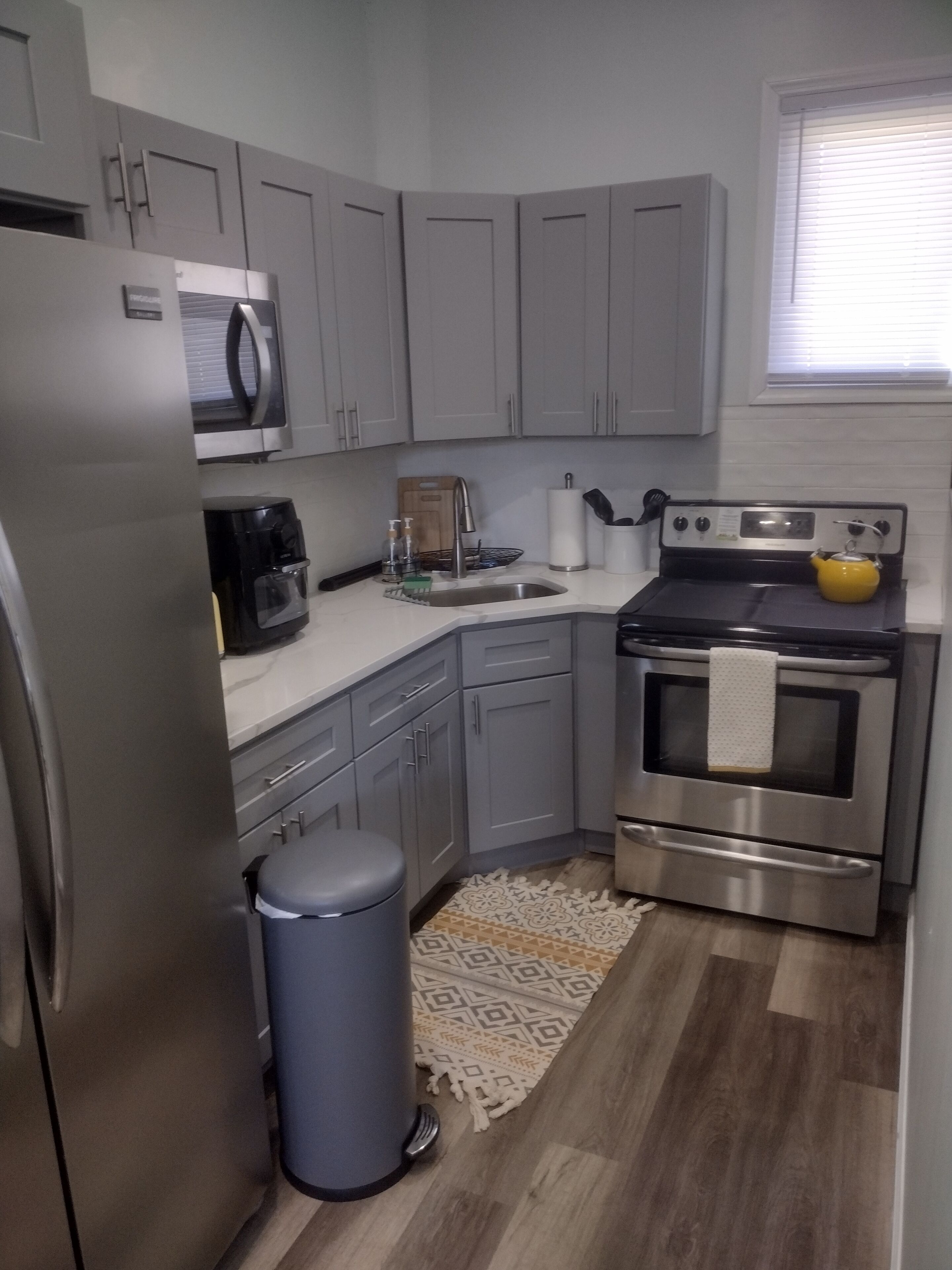 A compact kitchen with grey cabinets, a stainless steel refrigerator, a microwave, a coffee maker, a sink, a stove, and a trash bin.