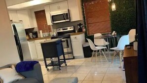 A modern living room with a sofa, ottoman, and a dining area with chairs and a table. A kitchen with appliances and a green wall is visible in the background. - Manatee Pocket! 1 Bedroom, 1 Bath Unit To Yourself (Stuart)