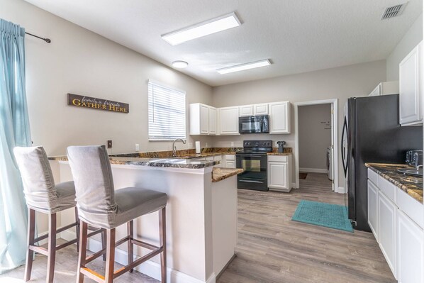 A modern kitchen with white cabinets, a black refrigerator, and a built-in oven. There are two bar stools and a window with blinds. - 185 Windwood Bay (Davenport)