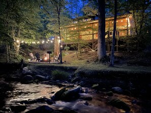 A riverside outdoor seating area with chairs and a fire pit, surrounded by trees and a building with string lights. - Creekside Firepit, Hot Tub, Serene Cabin Life!! (Gatlinburg)