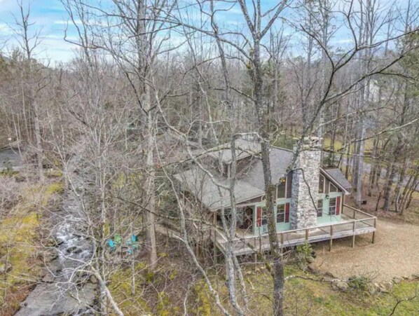 A cabin with a stone chimney, surrounded by trees and a small stream. - Creekside Firepit, Hot Tub, Serene Cabin Life!! (Gatlinburg)