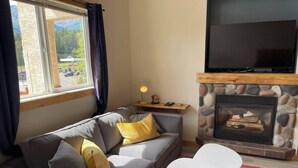 Ein Wohnzimmer mit Kamin aus Stein, einem grauen Sofa mit gelben Kissen, einem Flachbildfernseher über dem Kamin und einem roten Teppich mit Muster.