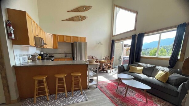 Eine moderne Küche mit Holzausstattung, einer zentralen Kochinsel mit Barhocker, einem Essbereich mit Tisch und Stühlen sowie einem Wohnzimmer mit einer Couch und einem großen Fenster mit Blick auf die Berge.