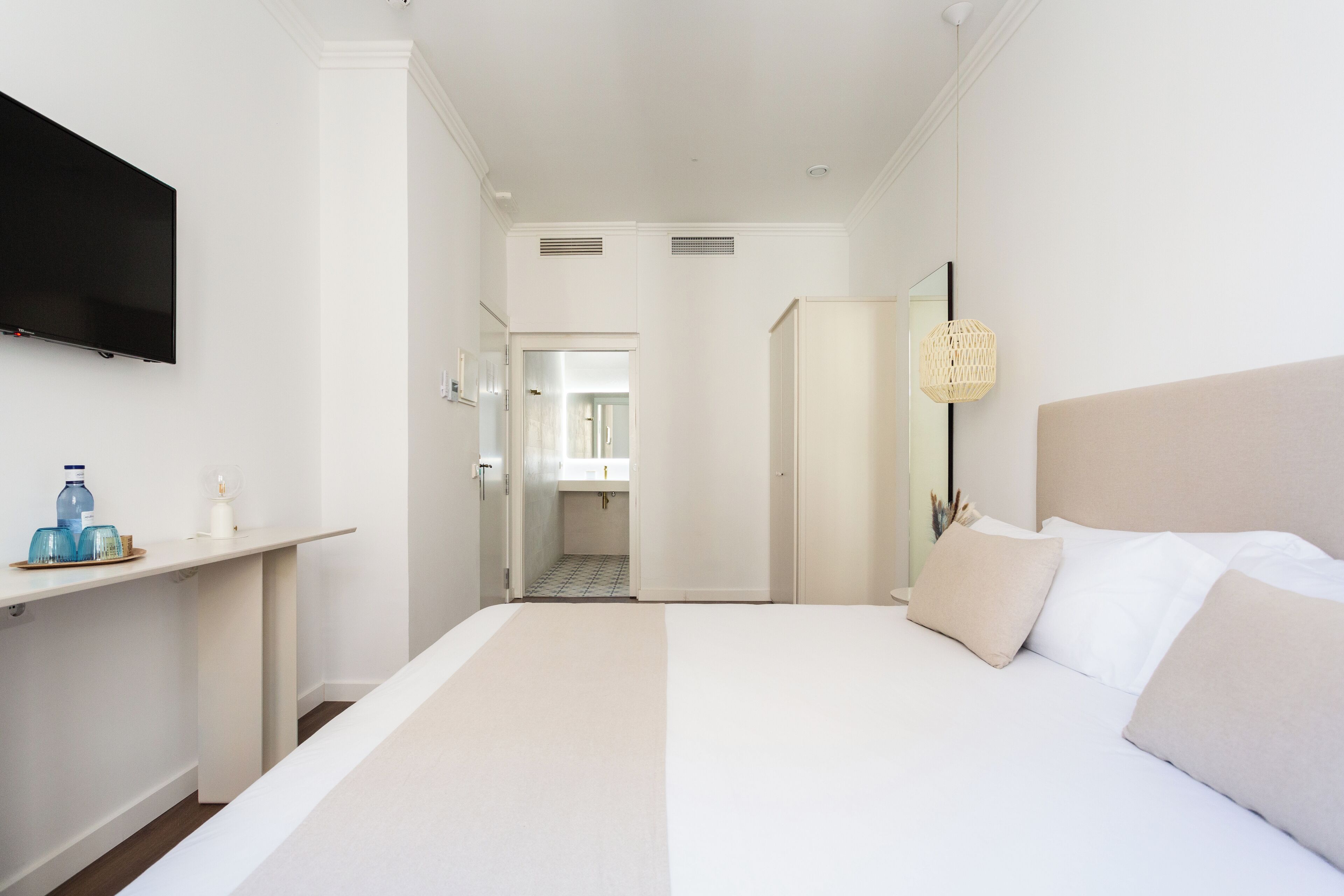 A hotel room with a large bed, a TV mounted on the wall, a bedside table with a water bottle, and a lamp.
