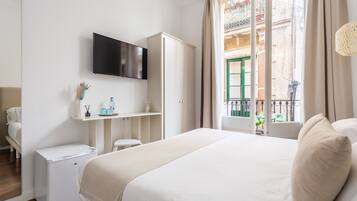 A hotel room with a bed, a TV mounted on the wall, a minibar, a small table, and a window with a view of a balcony.