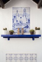 Un mur orné d’une mosaïque de carreaux bleus et blancs représentant une église, une étagère bleue avec des plantes en pot, et une rangée de carreaux décoratifs en dessous.